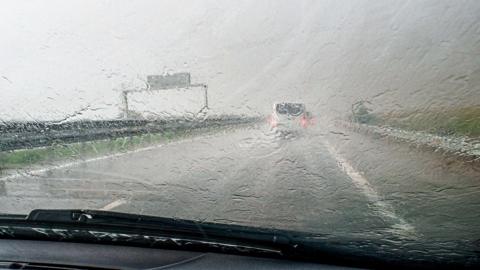 The view from a car window as it drives along a rainy, grey road. Cars ahead are barely visible due to the rain. 