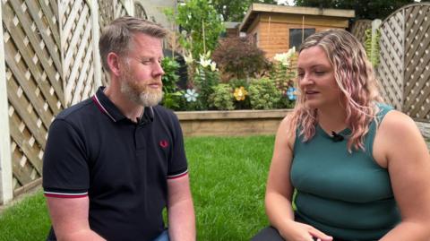 Dave and Charlotte Lay sitting in their garden. There is a small lawn behind them with fences either side of it and there are flowers and a garden shed at the end of the garden.