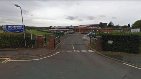 A Google street view image showing the entrance to a leisure centre car park. The green gates are open and a hedge is either side of the entrance. A sign on the left says "Wombourne Leisure Centre".