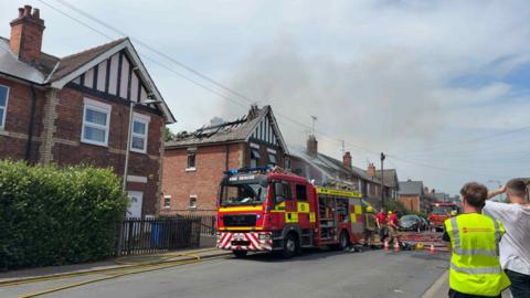 Fire engines outside a row of terraced houses. Smoke billows from above the houses.