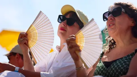 Two women in sunglasses fan themselves, with blue sky and sunshine overhead, while visiting London Zoo on 9 July.