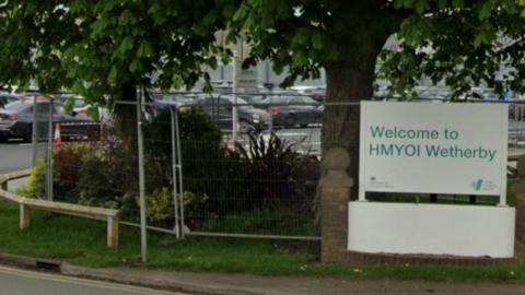 A white sign reading Welcome to HMYOI Wetherby on the side of a road. Behind the sign is a tree and a car park with lots of cars parked in it.