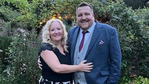 Lisa and Craig Shipley stand closely together with their arms around each other. They are smiling. Lisa has long, blonde, curly hair and she is wearing a black top with lace on the shoulders. Craig is wearing a grey suit with a purple tie. They are standing in a garden, in front of some bushes and shrubbery.