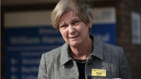 A blonde woman in a grey jacket and a yellow name tag with the name "Tracey" written on it.