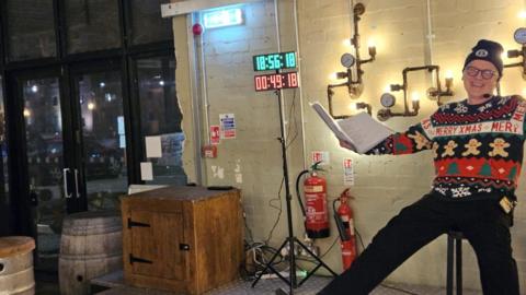 Dave Purchase sat back on a stool. He is wearing a blue, red, white and green christmas jumper - decorated with gingerbread men. He has on a blue hat and there is a large digital clock behind him. He is holding a lyrics book.