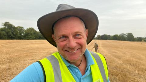 Gary Cook wearing a brimmed hat, high-vis jacket and light blue polo shirt, looking into the camera from a recently harvested field with detectorists in the background