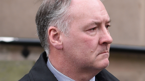 A close-up of a man's face from the side. A collar of a striped blue and white shirt and black jacket can be seen. The man has greying black hair cut in a short style, grey stubble and has a sombre expression on his face.