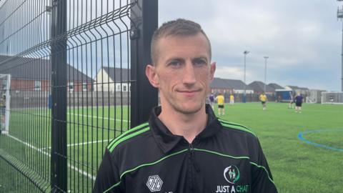 A man wearing a black and green "Just a Chat" training top is standing beside a fence of an outdoor football pitch, looking at the camera. Players are training in the background beneath an overcast sky