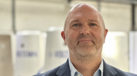 A man with a bald head and a faint beard with a grey jacket and white shirt in front of a blurred background