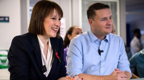 Rachel Reeves, wearing a suit jacket and a red poppy, stands to the right of Wes Streeting at a reception desk on a hospital ward. Medical staff stand behind them. They are both clasping their hands in front of them and they are smiling at the staff member behind the desk