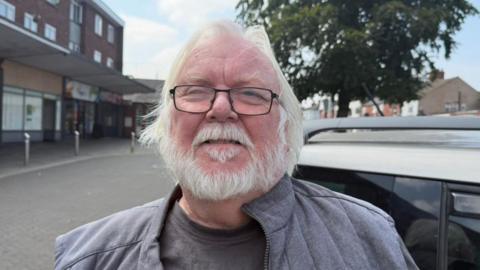 The photo shows Rob Barnard looking at the camera. He has white hair and a white beard, and is wearing black rimmed glasses. He's also wearing a grey jacket. He's standing in front of a car. Behind him, to his left, there are some shops.