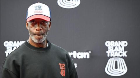 Michael Johnson looking pensive as he stands in front of a black board bearing the Grand Slam Track name and logo