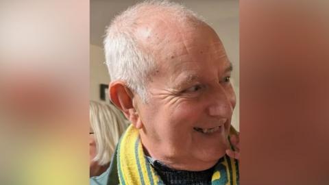 Jim Davies is pictured smiling. He is balding, with some short, white hair and is wearing a yellow and green football scarf.