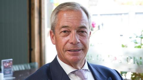 Nigel Farage sitting in a pub while wearing a white shirt and navy blazer. He is smiling.