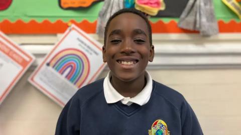 Easton smiling at the camera. He has short dark hair and is wearing a white collared shirt under a navy blue sweatshirt with the school logo on the chest. 