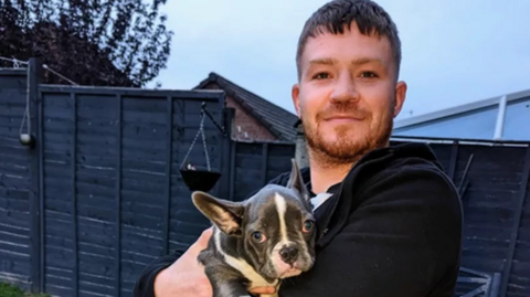 Ben Daly has brown short hair and is wearing a black hoodie. He is holding a dog while standing in his back garden. Behind him are black fence panels and the sky is cloudy. 