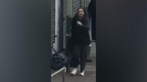 A blurry CCTV image of a woman in black trousers and a black jacket walking down the street. She is wearing white trainers. 