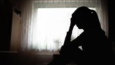 A woman sat in front of a window and in shadow, with her hand on her forehead. 