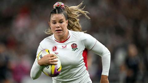 Jess Breach is wearing a white England jersey. She is running with a rugby ball. 