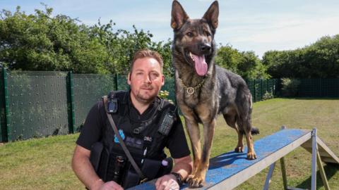 A man in police uniform next to Jura, a German Shepherd wearing a gold chain with his tongue out.