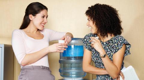 Two woman either side of a blue watercooler, both holding white plastic cups. Both have long dark hair, the one on the left has short hair, the one on the right has longer curly hair.  The woman on the left has a pink top while the one on the right has a black and white dress and is holding some papers.  They are both smiling.