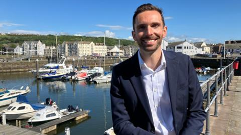 Josh MacAlister, who is the Member of Parliament for Whitehaven and Workington in the North West of England, smiles for a photograph in front of a harbour. 