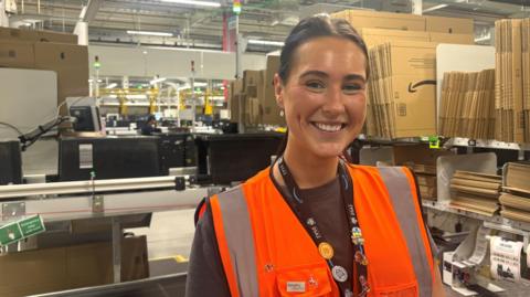 A head and shoulders shot of Grace Rutter smiling at the camera. She is wearing a black t-shirt, an orange hi-vis vest and a black Amazon lanyard with badges on. She has long, dark brown hair which is tied back. She is standing in a warehouse. Cardboard boxes, a conveyer belt and machinery are behind her. 