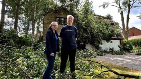 Rosamund and Richard Booth stand outside their home which is partially obscured by the branches of a tree that has fallen on the roof of their house. There are also numerous branches lying in their garden and at their feet