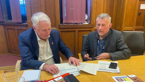 Council leader George Duggins and finance cabinet member Richard Brown sit at a desk in a wood-panelled room, looking over tables of figures and other paperwork. Both men are wearing suit jackets with open-necked shirts. 