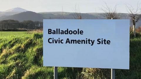 A large white sign with the word Balladoole Civic Amenity Site written on it in black lettering, in a grassed area with green hills in the background.