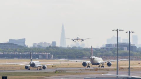 Two planes driving away from the camera on a runway at Heathrow Airport, with buildings including the Shard in the background. Another plane is facing the camera and coming in to land