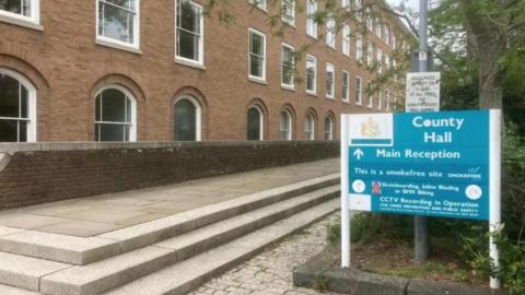 A general view of Devon County Hall. It shows a sign that says County Hall while there is a brown brick building in the background.