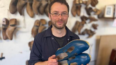 A man in a navy boiler suit with short dark hair and glasses holds up a pair of blue clogs.