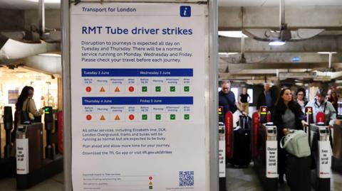 In the foreground, a board carrying a poster titled "RMT Tube drivers strikes" with warning of disruption and schema of when it will impact travel. In the background, to the left, are people walking past TfL barriers at a tube station