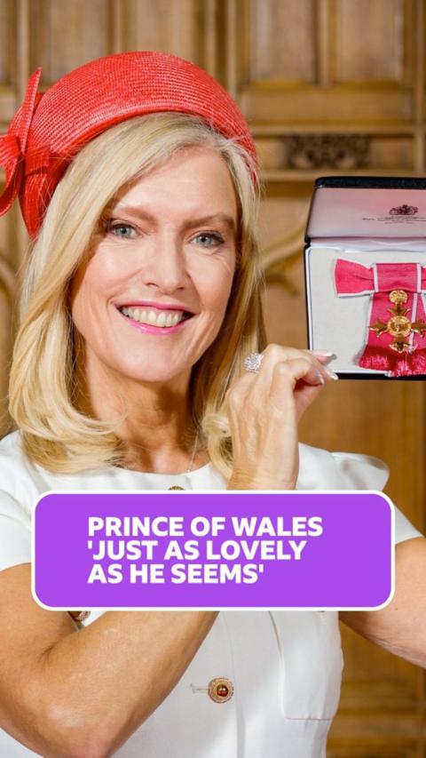 Linda Magistris is smiling at the camera holding up her OBE. She has mid length blonde hair with an orange hat and white smart short sleeved dress with gold button detailing