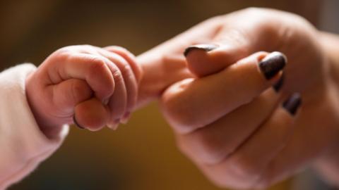 A new baby holds the finger of their mother whose hand has painted nails. The background is blurred. 