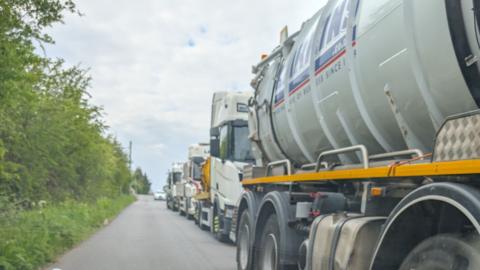 Large white and yellow tankers line up on Lowfield Road in Bolton Upon Dearne to take wastewater away. A white car is at the back of the queue with trees to the left of the tarmac.