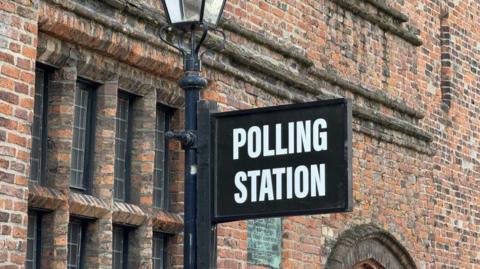 A black sign reading 'POLLING STATION' is attached on a Victorian-style lamp post, which is in front of a red-brick period property. Only a section of the building is visible: the top half of a window.