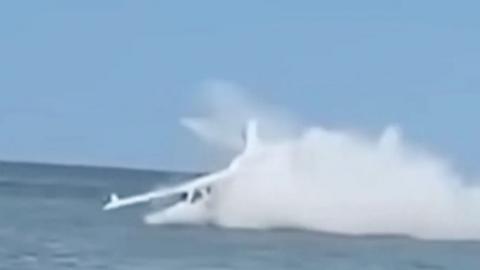 Still of a white personal aircraft crash landing into water. Jet spray is flying up around the plane.