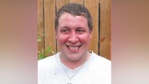 Steven Peck. He has short light brown hair, wearing a white t-shirt and silver chain with a cross.