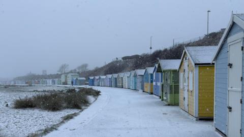 A row of beach huts along a curved path. They are painted different colours, and there is snow settled on the walkway in front of them.