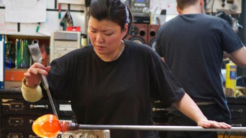 A woman creates a jar at a glass-blowing factory. The jar is glowing red at the end of the pole used to create it.