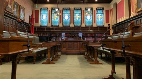 An empty House of Keys chamber with dark wooden benches and green-leather chairs. It is a dark wooden-panelled room overseen by six stained glass windows.