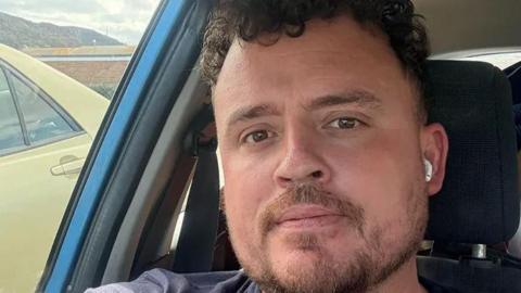 Guy Bradshaw with brown eyes, curly hair and beard and moustache wearing a blue t-shirt sitting in a blue vehicle. He has white headphones in. He is parked alongside a yellow car with mountains in the background. 
