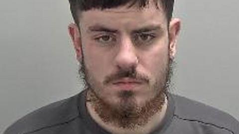 Frankie Maughan in his mugshot photo. It is low quality, he has a beard and is stood against a plain background. 