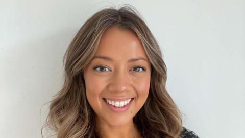 Victoria Cleave smiles at the camera. She has light brown hair with some blonde highlights through it. She stands against a white wall.