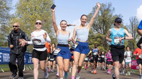 Two women in matching grey tops and blue shorts running while holding hands, with their other hands in the air. A man in a jacket, a smiling woman in a white top, and a man in a cap and blue tank top are running to their sides. dozens of other runners are seen in the background.
