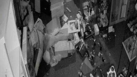 A black and white CCT image shows a male in a hooded top sprawled across the floor after falling over in an antiques shop. He has tripped over a pile of boxes and is surrounded by broken items.