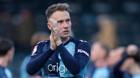 Cauley Woodrow, wearing the light and dark blue quarters of Wycombe Wanderers, claps the fans