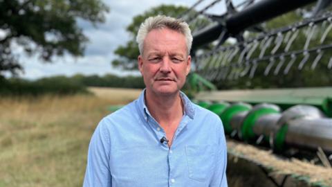 The National Farmers' Union's deputy president looking at camera with a serious expression. He has grey short hair and a blue shirt. He is standing in front of farming equipment in one of his fields.